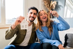 Casal jovem sorridente sentado no sofá celebrando a casa nova, com o homem segurando uma miniatura de casa branca e a mulher mostrando as chaves.
