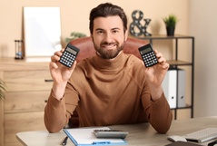 Homem sorridente sentado em escritório, segurando dois calculadoras, com papel e caneta na mesa, simbolizando sucesso financeiro.