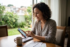 Mulher sorridente navega no smartphone à mesa de casa, com jardim verde ao fundo e café ao lado, transmitindo tranquilidade ao gerenciar suas finanças digitalmente.