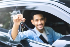 Homem sorridente sentado no banco do motorista de um carro preto segurando as chaves do veículo para fora da janela.
