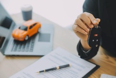 Mão segurando a chave de um carro sobre uma mesa com contrato assinado e uma miniatura de veículo laranja ao fundo.