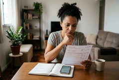 Mulher lê um documento com expressão séria e pensativa à mesa da sala, com caderno e smartphone ao lado, refletindo sobre uma situação financeira que exige atenção.