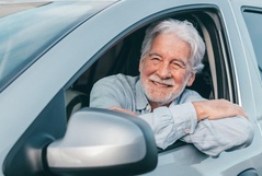 Homem idoso sorridente com cabelos brancos apoiado na janela aberta do banco do motorista de um carro prata.