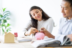 Casal sorridente colocando uma moeda em um cofrinho de porquinho rosa sobre uma mesa com uma maquete de casa ao lado.
