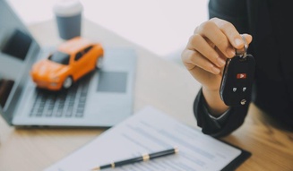 Mão segurando a chave de um carro sobre uma mesa com contrato assinado e uma miniatura de veículo laranja ao fundo.