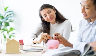 Casal sorridente colocando uma moeda em um cofrinho de porquinho rosa sobre uma mesa com uma maquete de casa ao lado.