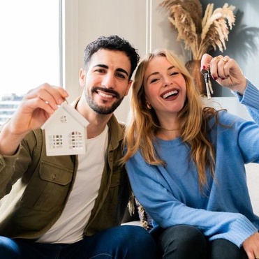 Casal jovem sorridente sentado no sofá celebrando a casa nova, com o homem segurando uma miniatura de casa branca e a mulher mostrando as chaves.