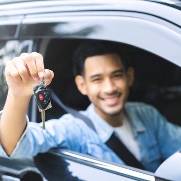 Homem sorridente sentado no banco do motorista de um carro preto segurando as chaves do veículo para fora da janela.