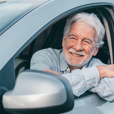 Homem idoso sorridente com cabelos brancos apoiado na janela aberta do banco do motorista de um carro prata.