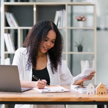 Mulher sorridente organizando finanças em um escritório, usando uma calculadora e escrevendo em papéis.