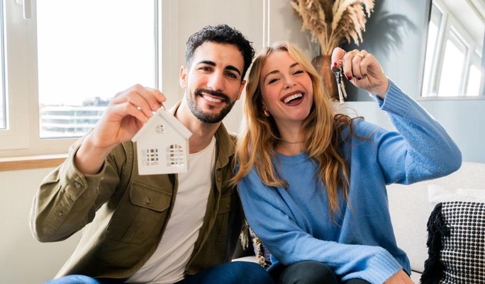 Casal jovem sorridente sentado no sofá celebrando a casa nova, com o homem segurando uma miniatura de casa branca e a mulher mostrando as chaves.
