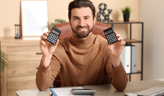 Homem sorridente sentado em escritório, segurando dois calculadoras, com papel e caneta na mesa, simbolizando sucesso financeiro.