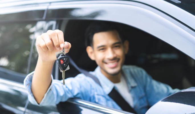 Homem sorridente sentado no banco do motorista de um carro preto segurando as chaves do veículo para fora da janela.