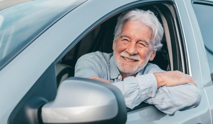 Homem idoso sorridente com cabelos brancos apoiado na janela aberta do banco do motorista de um carro prata.