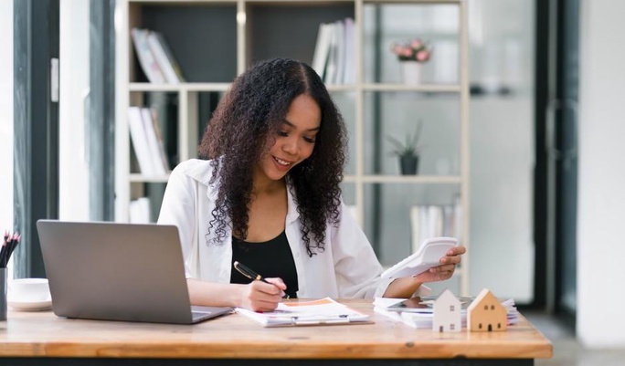 Mulher sorridente organizando finanças em um escritório, usando uma calculadora e escrevendo em papéis.